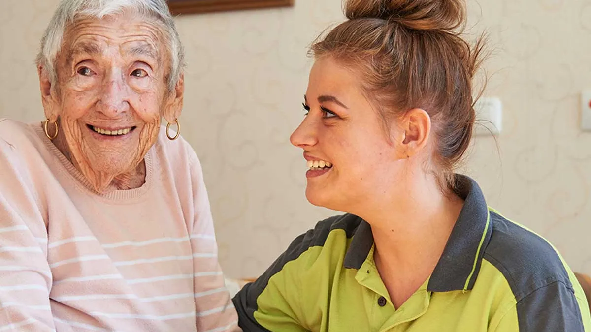 Pflegerin blickt lächelnd zu einer ebenfalls lächelnden Bewohnerin von PFLEGEN & WOHNEN HAMBURG Marienthal.