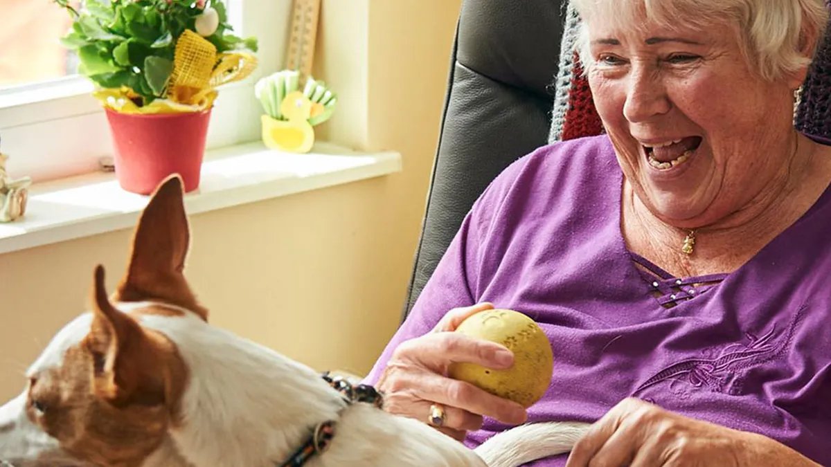 Bewohnerin von PFLEGEN & WOHNEN HAMBURG in Heimfeld spielt lachend mit dem Hund auf ihrem Schoß und hält einen Ball.
