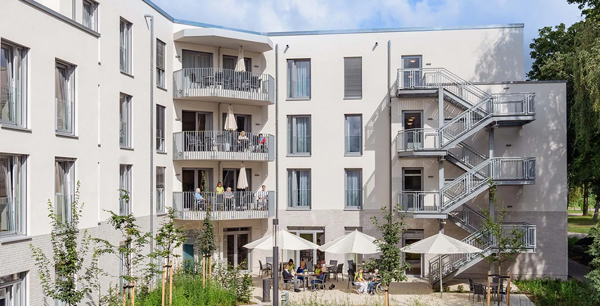 Außenansicht von PFLEGEN & WOHNEN HAMBURG Marienthal mit Gemeinschaftsterrasse, Sitzmöglichkeiten und Balkone in grüner Lage.