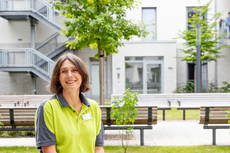 Die Pflegefachkraft Aida Bahic steht vor dem modernen Gebäude von PFLEGEN & WOHNEN MARIENTHAL und trägt ein grünes Arbeitsoberteil.