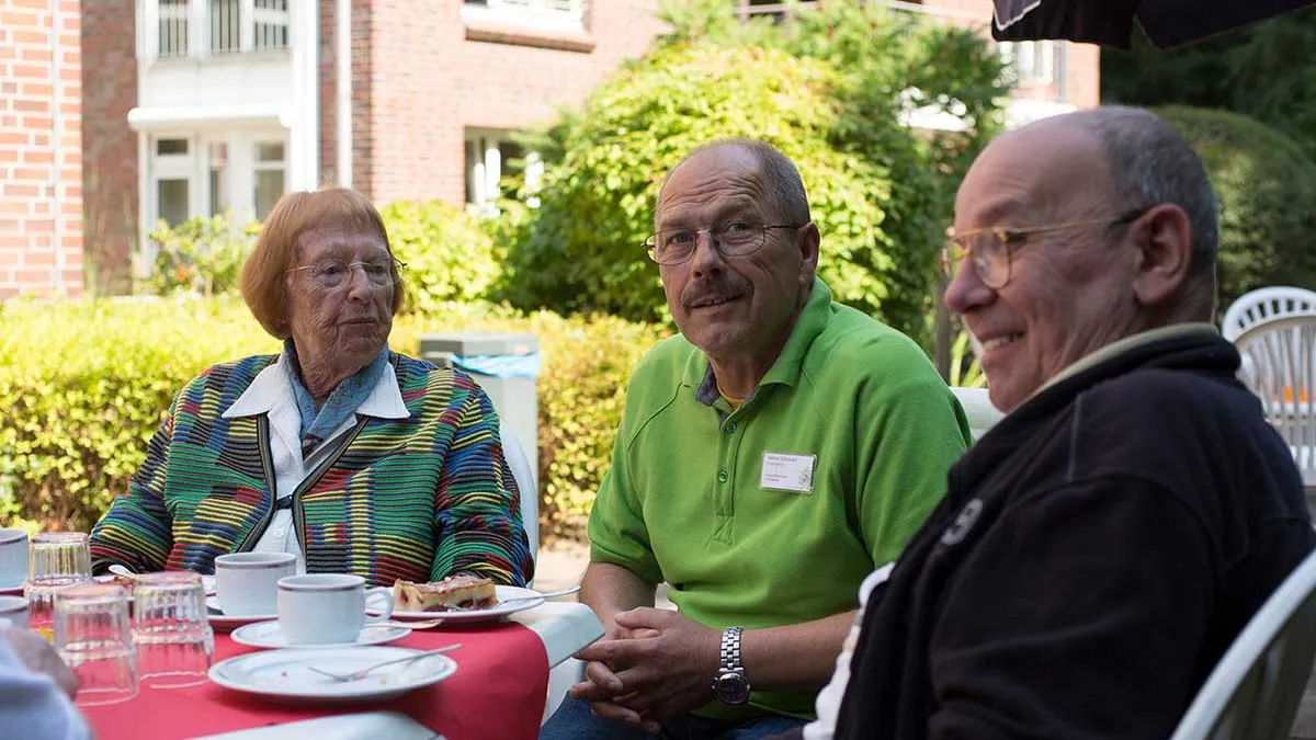 Pfleger sitzt draußen zusammen mit Bewohnern von PFLEGEN & WOHNEN HAMBURG am Standort Lutherpark bei Kaffee und Kuchen.