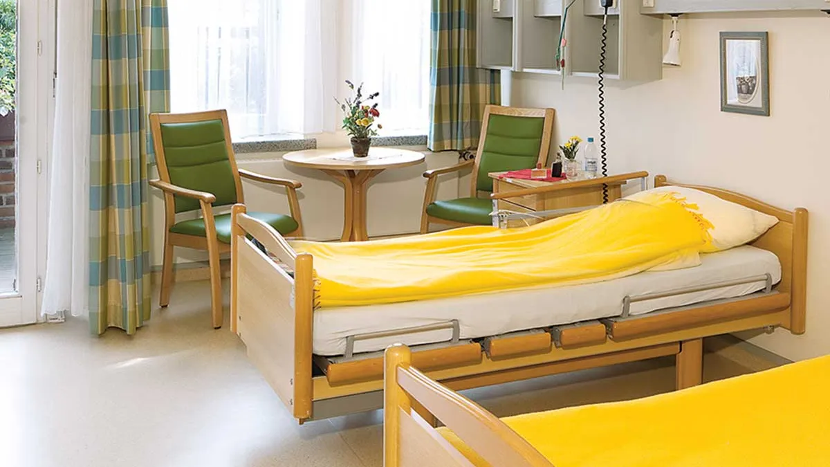 Raum mit zwei Betten im PFLEGEN & WOHNEN HAMBURG in Altona. Neben einem Bett steht ein Nachttisch. Daneben ist am Fenster eine Sitzgruppe.