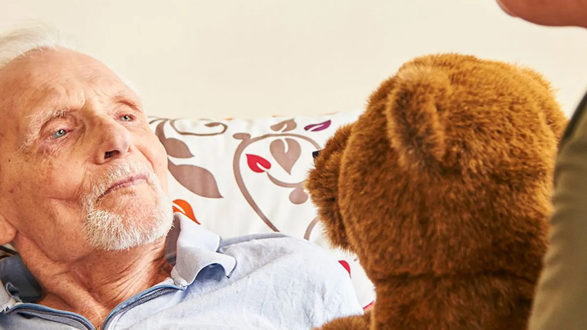 Mitarbeiterin überreicht einem Bewohner von PFLEGEN & WOHNEN HAMBURG in Heimfeld einen Teddybären.