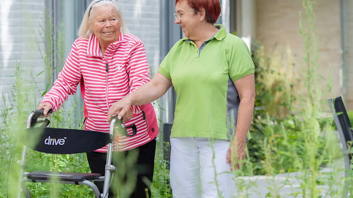Bewohnerin mit Rollator geht zusammen mit Pflegerin am PFLEGEN & WOHNEN HAMBURG-Standort Marienthal spazieren.